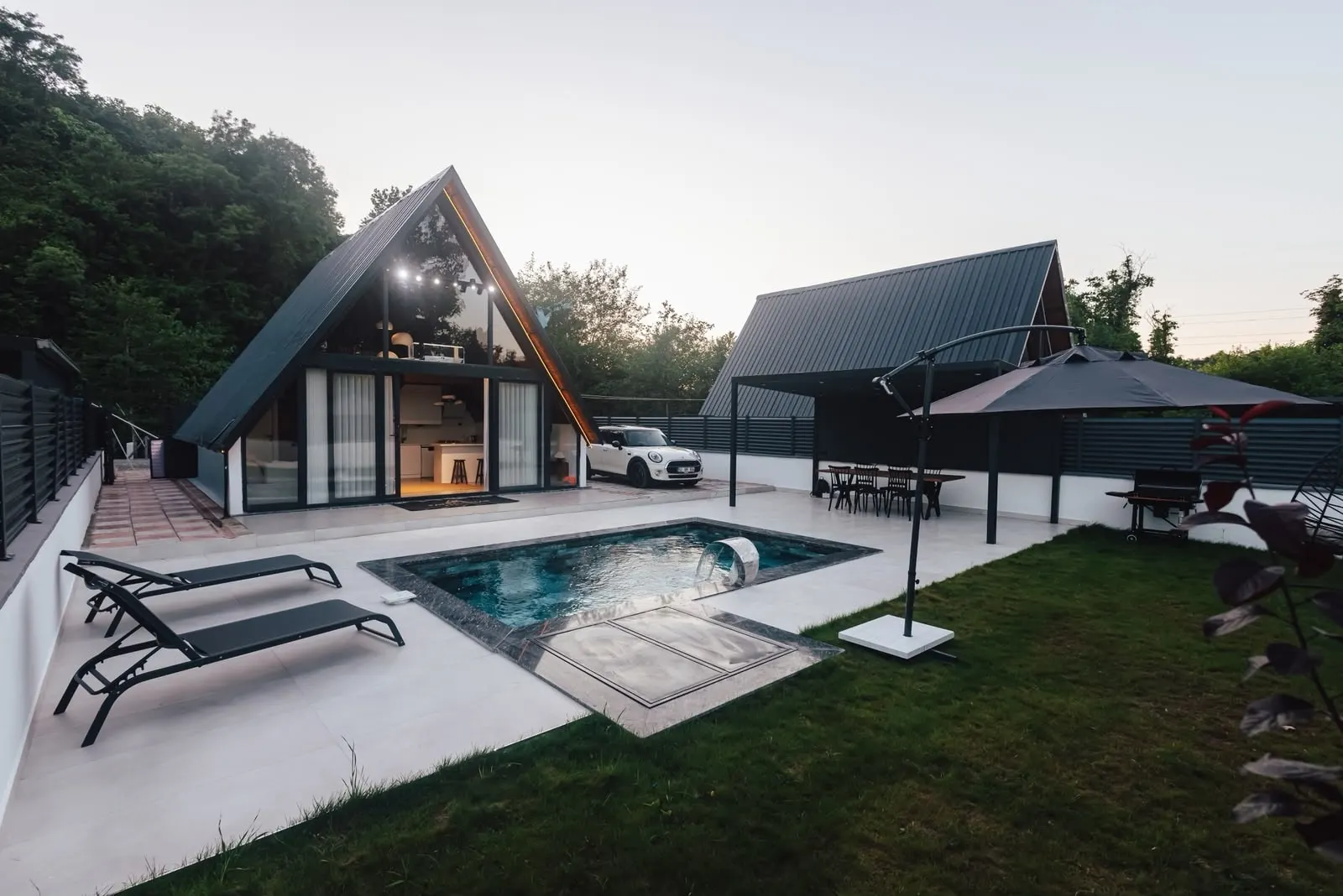 Irmak bungalovunun çim bahçesi, şezlonglar ve havuz; arkada A-ev ve pergola